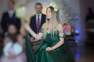 Tânără în rochie verde cu coroană florală și microfon headset, decor floral și un copil blurat în prim-plan