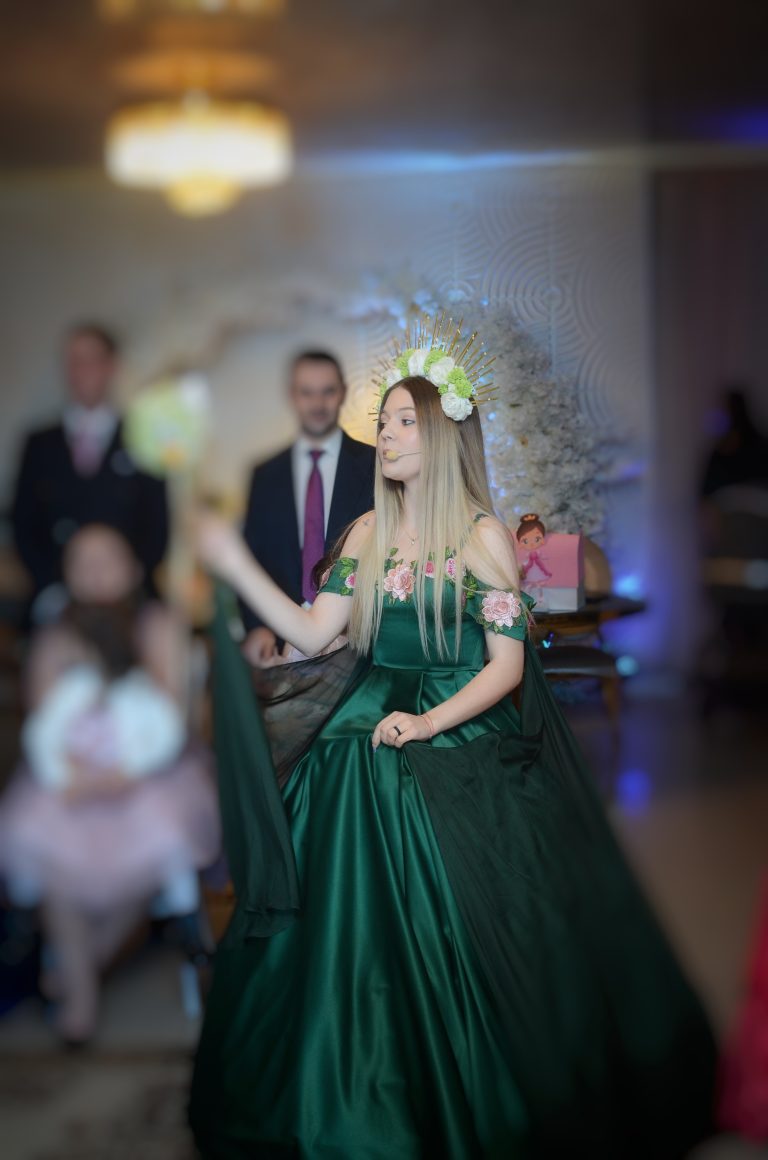Tânără în rochie verde cu coroană florală și microfon headset, decor floral și un copil blurat în prim-plan