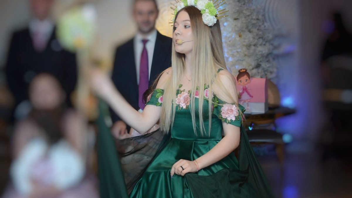 Tânără în rochie verde cu coroană florală și microfon headset, decor floral și un copil blurat în prim-plan