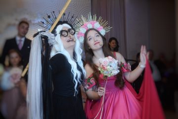 Două persoane costumate pe scenă, una în negru cu perucă albă și coroană cu țepi, cealaltă în rochie roz cu cunună de flori, țin microfoane headset