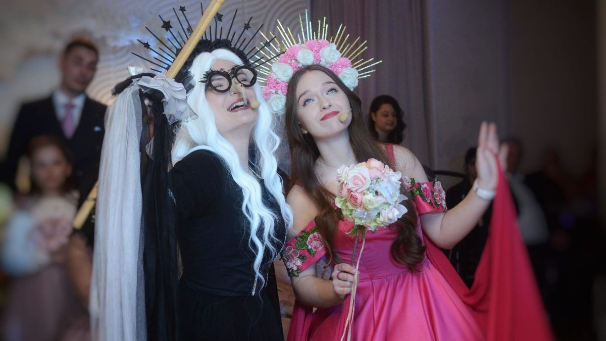 Două persoane costumate pe scenă, una în negru cu perucă albă și coroană cu țepi, cealaltă în rochie roz cu cunună de flori, țin microfoane headset