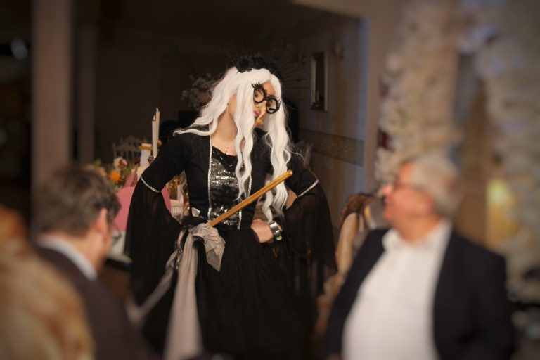 Persoană îmbrăcată ca animator costumat pentru petrecere, cu perucă albă lungă, ochelari supradimensionați și recuzită de lemn, în interior la mese decorate.