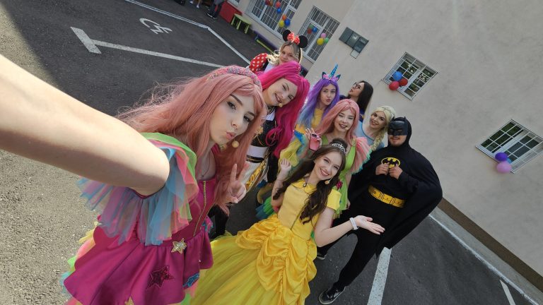 Grup de animatori costumați — prințese, unicorn, Minnie și Batman — pozând pentru selfie, cu baloane în fundal