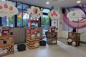 Interior cu standuri din lemn etichetate «Vată de zahăr», «Popcorn» și «Punct de încasare», mașină de popcorn, echipamente pentru vată de zahăr și decorațiuni tematice.