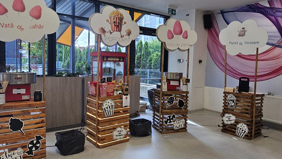 Interior cu standuri din lemn etichetate «Vată de zahăr», «Popcorn» și «Punct de încasare», mașină de popcorn, echipamente pentru vată de zahăr și decorațiuni tematice.