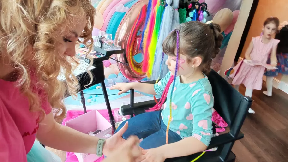O adultă aplică pictură pe față și prinde extensii colorate unui copil la Beauty Bar copii, masa cu palete și extensii colorate în fundal