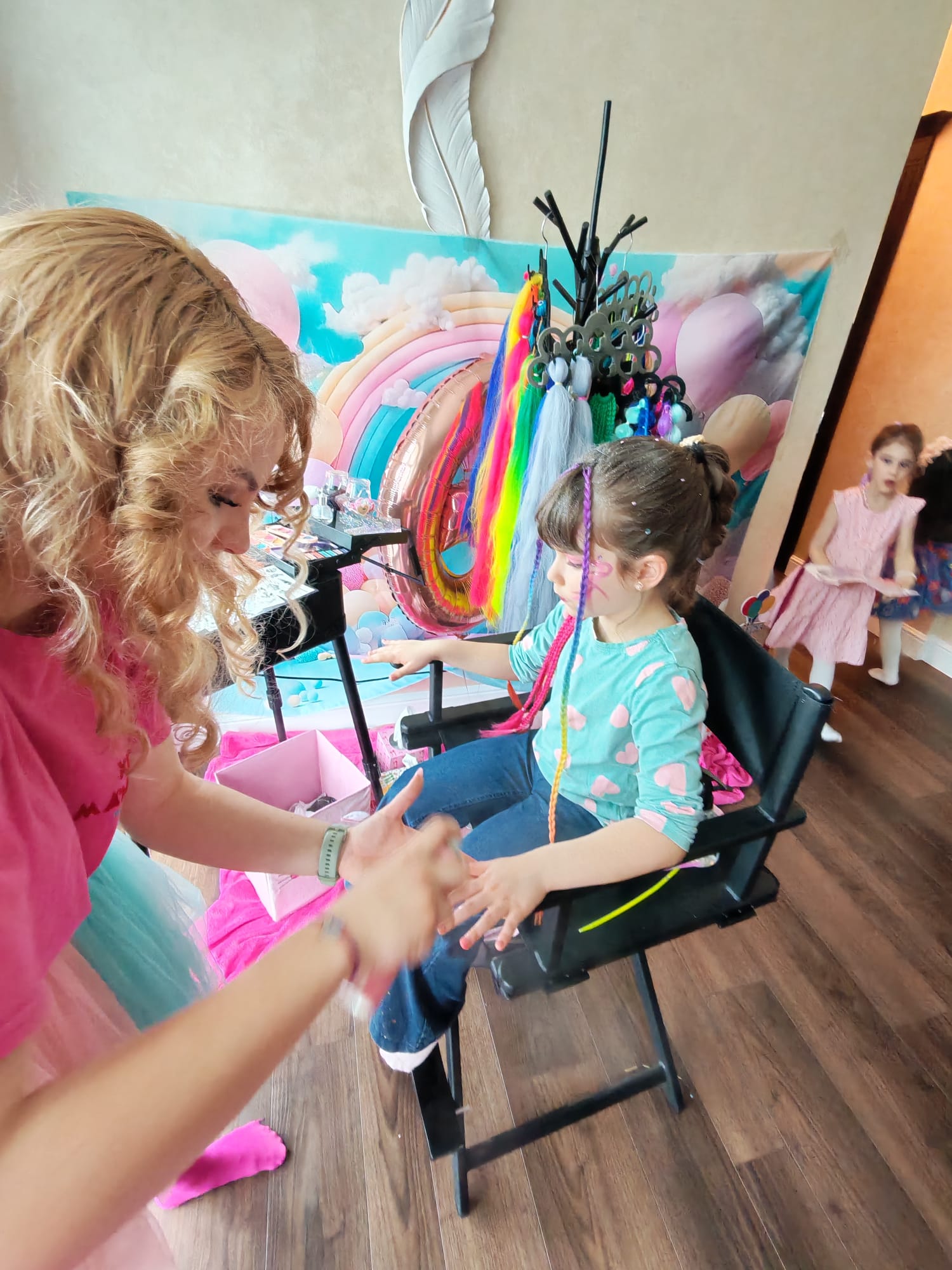 O adultă aplică pictură pe față și prinde extensii colorate unui copil la Beauty Bar copii, masa cu palete și extensii colorate în fundal