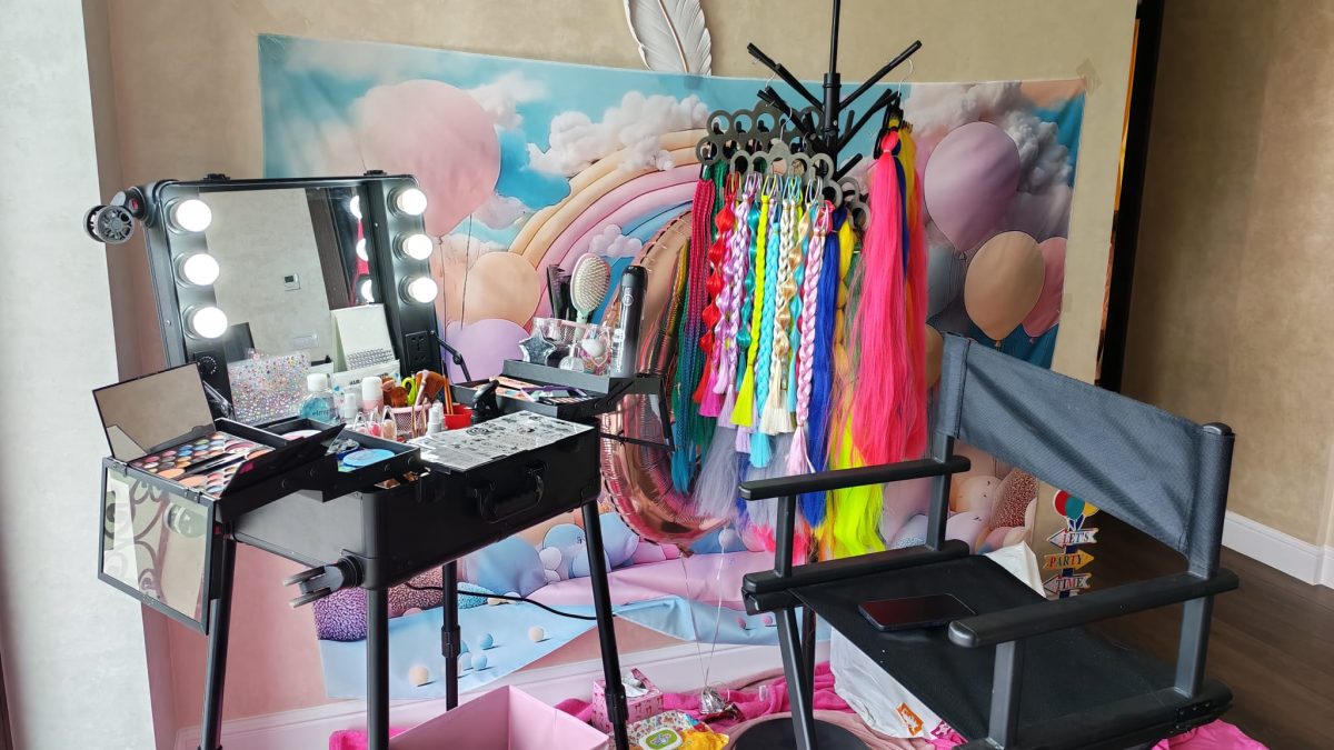 Stație de machiaj iluminată cu palete, pensule și cozi de păr colorate lângă baloane și cutii textile.