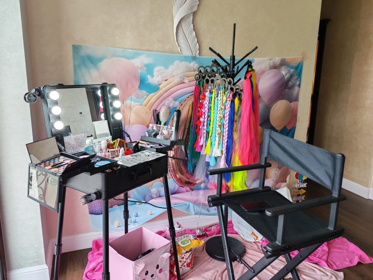 Stație de machiaj iluminată cu palete, pensule și cozi de păr colorate lângă baloane și cutii textile.