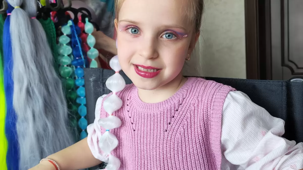 Fetiță cu machiaj roz și extensie albă împletită, accesorii colorate și o persoană în fundal aranjând părul.