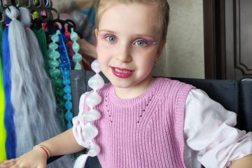 Fetiță cu machiaj roz și extensie albă împletită, accesorii colorate și o persoană în fundal aranjând părul.