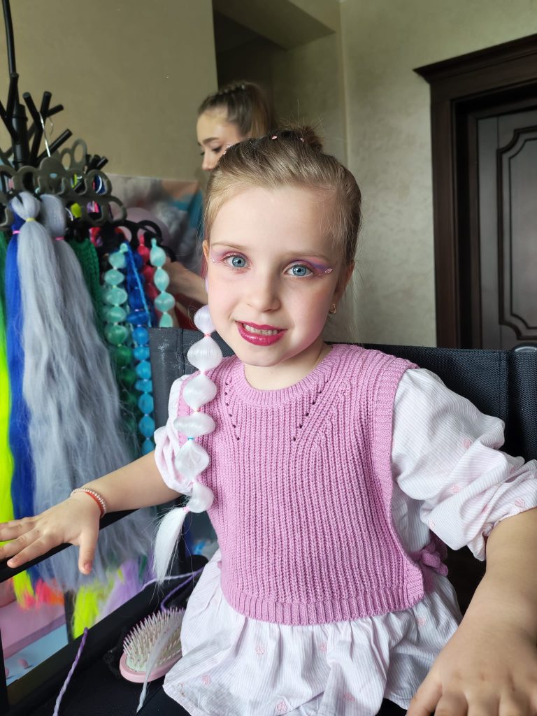 Fetiță cu machiaj roz și extensie albă împletită, accesorii colorate și o persoană în fundal aranjând părul.