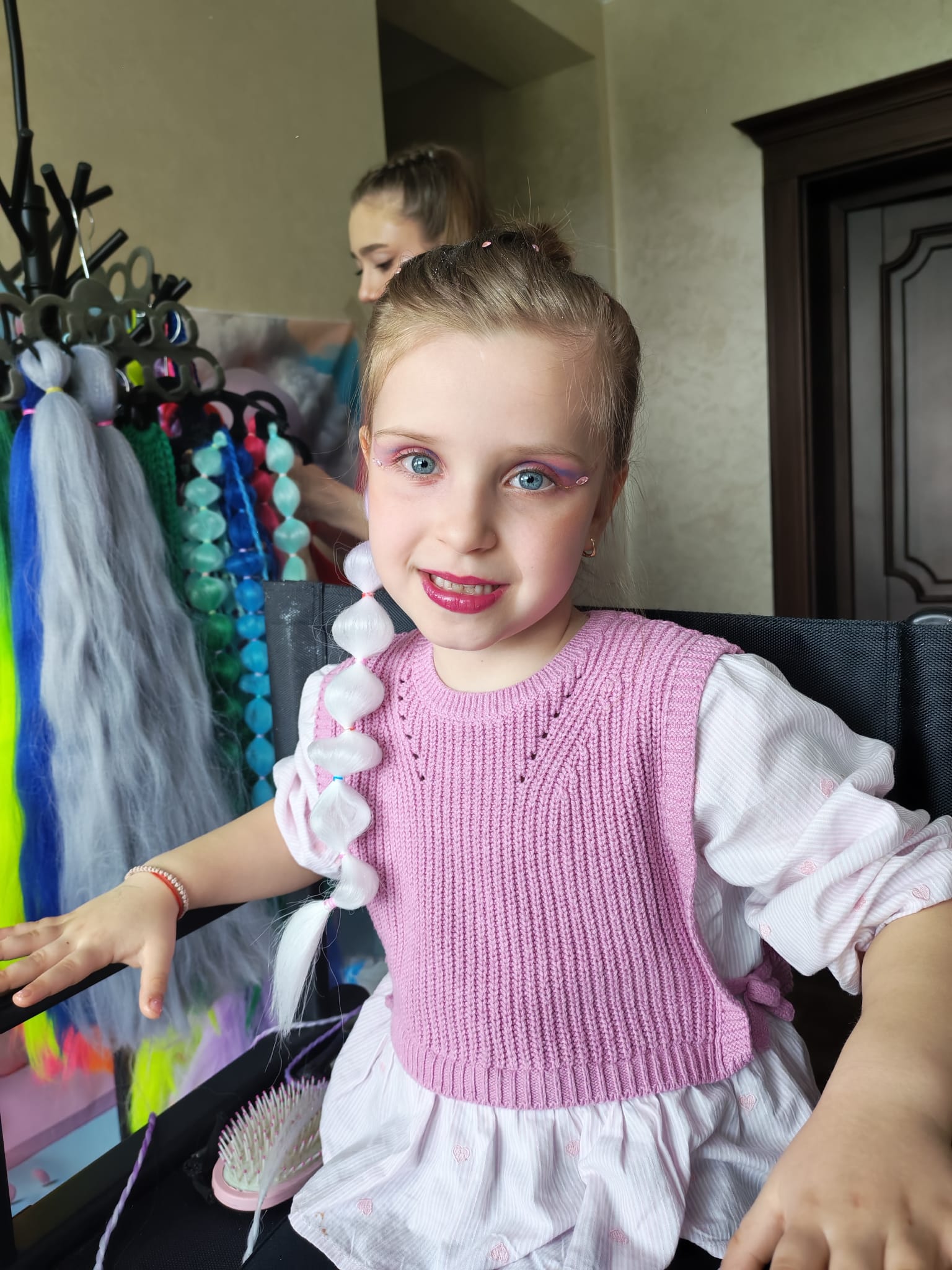 Fetiță cu machiaj roz și extensie albă împletită, accesorii colorate și o persoană în fundal aranjând părul.
