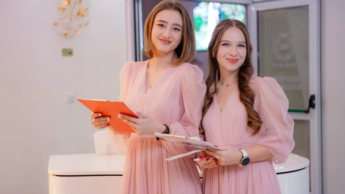 Două femei în rochii roz zâmbesc ținând clipboard-uri într-un interior cu ușă deschisă