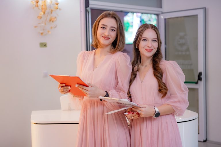 Două femei în rochii roz zâmbesc ținând clipboard-uri într-un interior cu ușă deschisă