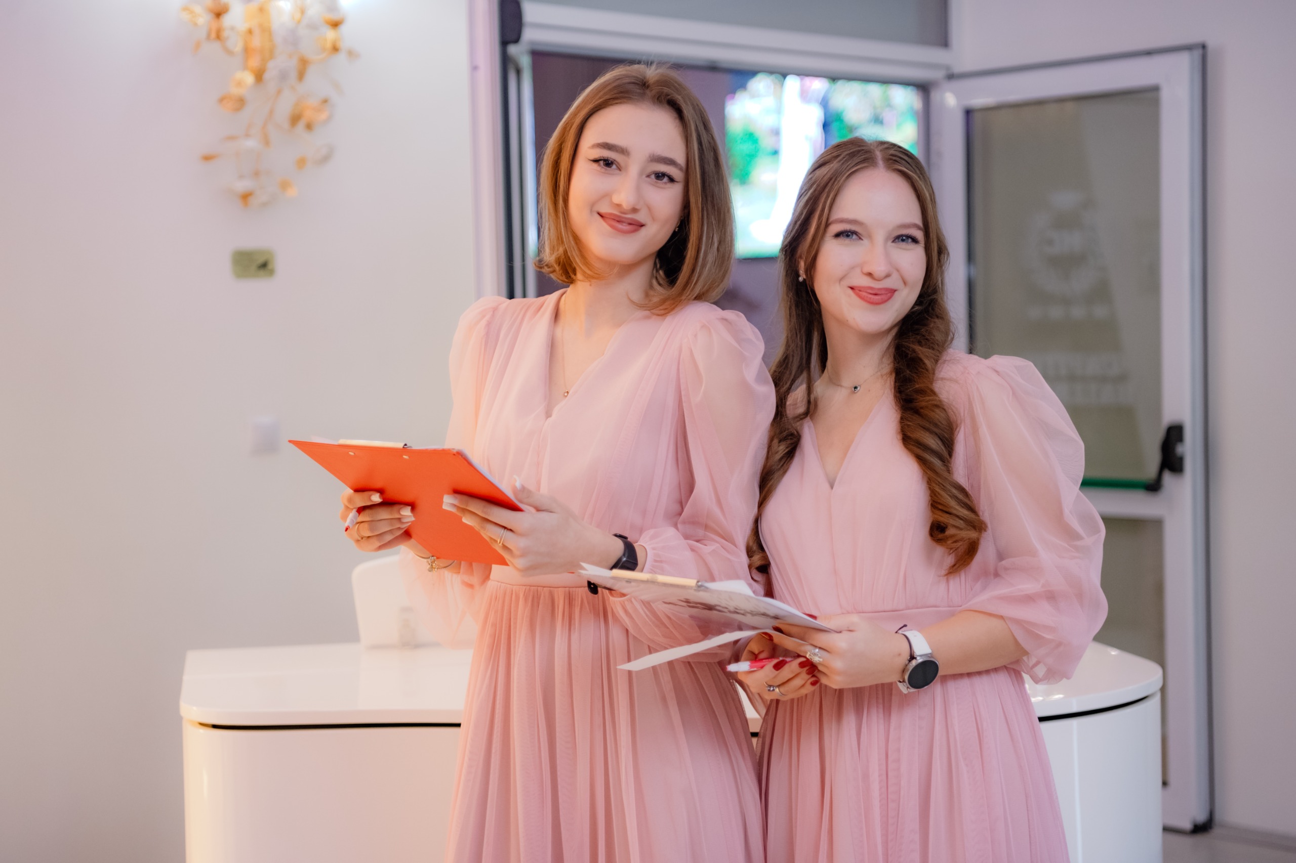 Două femei în rochii roz zâmbesc ținând clipboard-uri într-un interior cu ușă deschisă