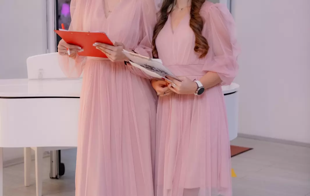 Două hostess îmbrăcate în rochii roz, ținând clipboarduri lângă un pian alb, imagine de hostess la petrecere pentru copil