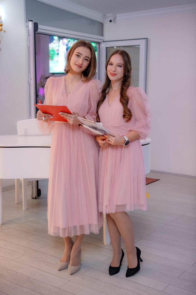 Două hostess îmbrăcate în rochii roz, ținând clipboarduri lângă un pian alb, imagine de hostess la petrecere pentru copil