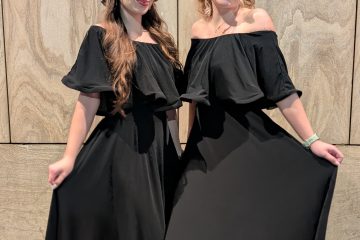Două persoane în rochii negre zâmbind în fața unui perete de piatră, atmosferă festivă