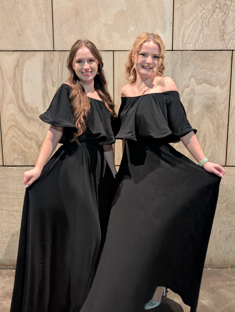 Două femei elegante în rochii negre stând una lângă alta în fața unui perete din piatră