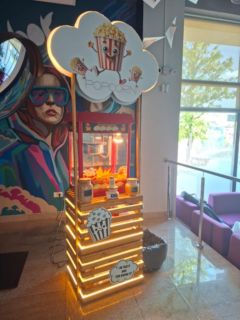 Stand de popcorn iluminat lângă o pictură murală colorată și zone de relaxare.