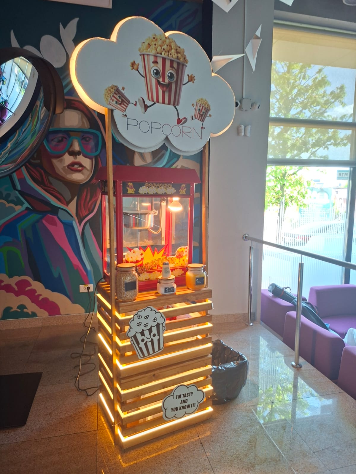 Stand de popcorn iluminat lângă o pictură murală colorată și zone de relaxare.