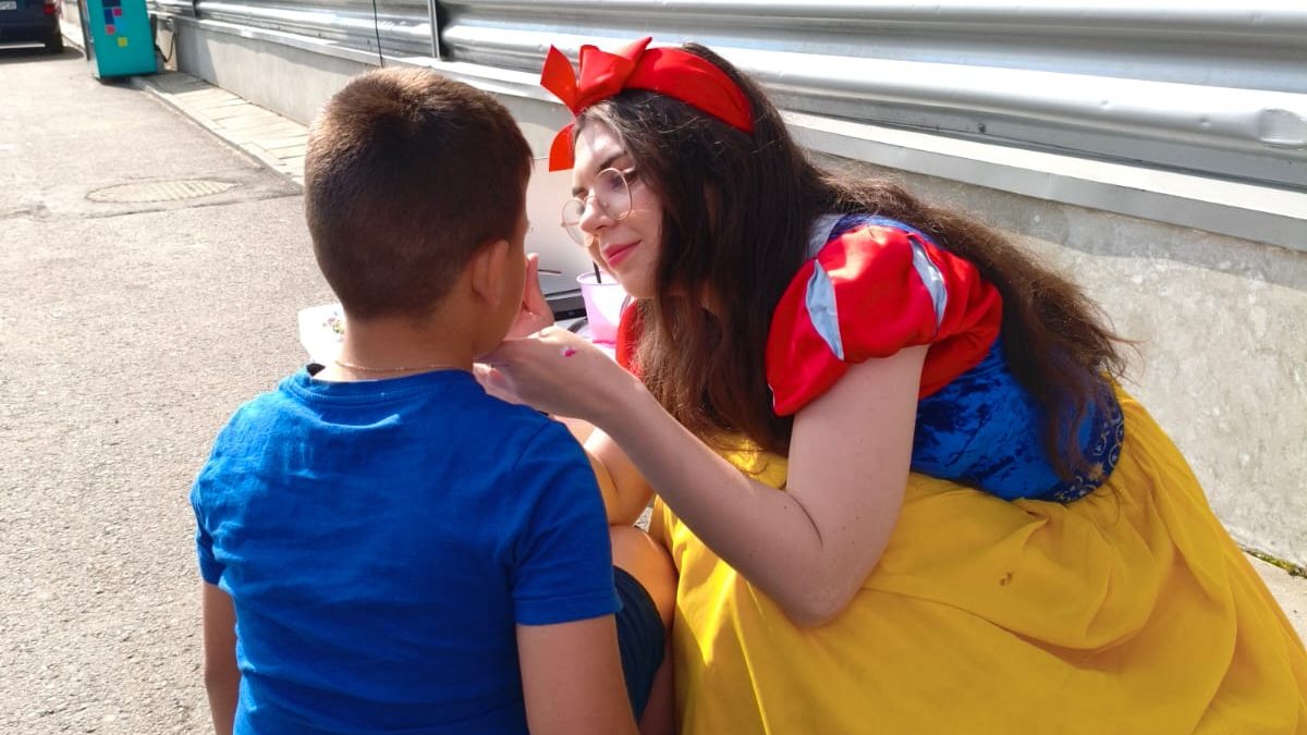 Adult îmbrăcat ca Snow White aplică pictură pe fața unui copil pe scăunel; animatorilor pentru pictură în acțiune, pictură pe față într-un decor outdoor