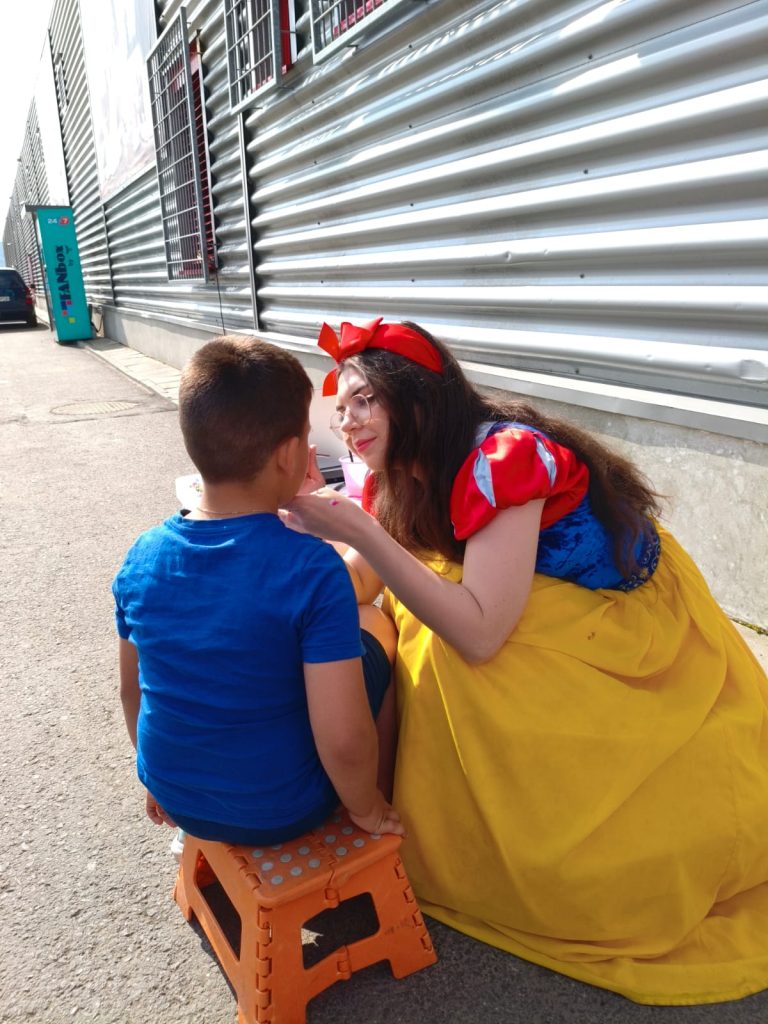 Adult îmbrăcat ca Snow White aplică pictură pe fața unui copil pe scăunel; animatorilor pentru pictură în acțiune, pictură pe față într-un decor outdoor